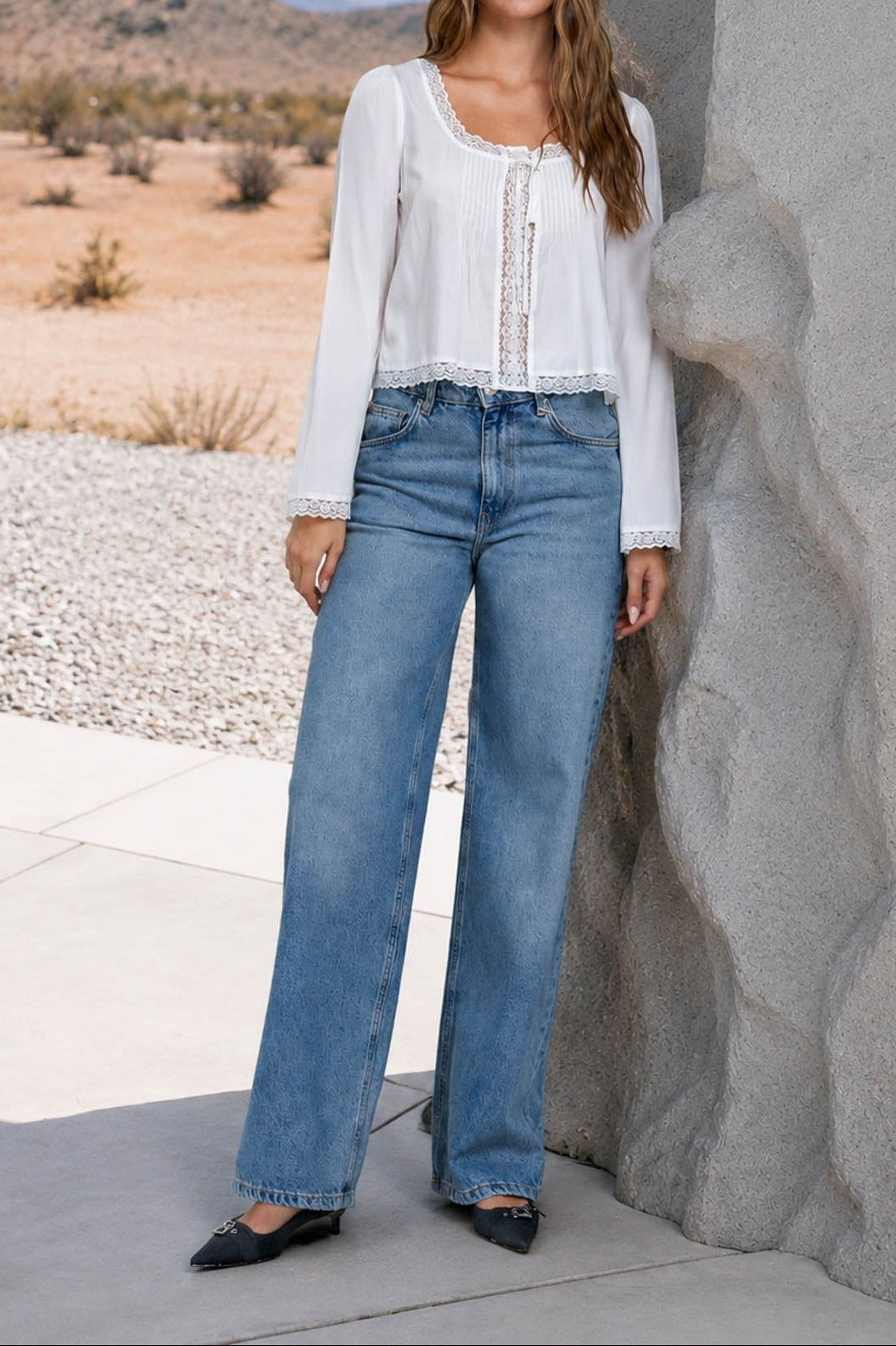 A woman stands outdoors against a large rock formation wearing a white long-sleeve blouse with lace-up front detailing and wide-leg blue jeans, with a desert landscape in the background.