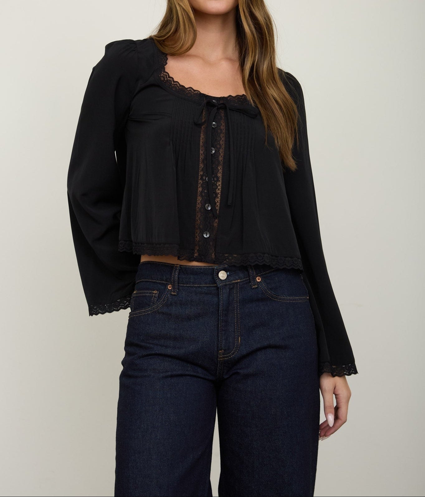 A woman modeling a black long-sleeve blouse with lace trim and a pleated front, paired with dark blue jeans, against a plain light gray studio background.