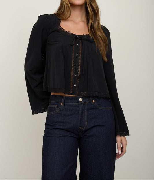 A woman modeling a black long-sleeve blouse with lace trim and a pleated front, paired with dark blue jeans, against a plain light gray studio background.