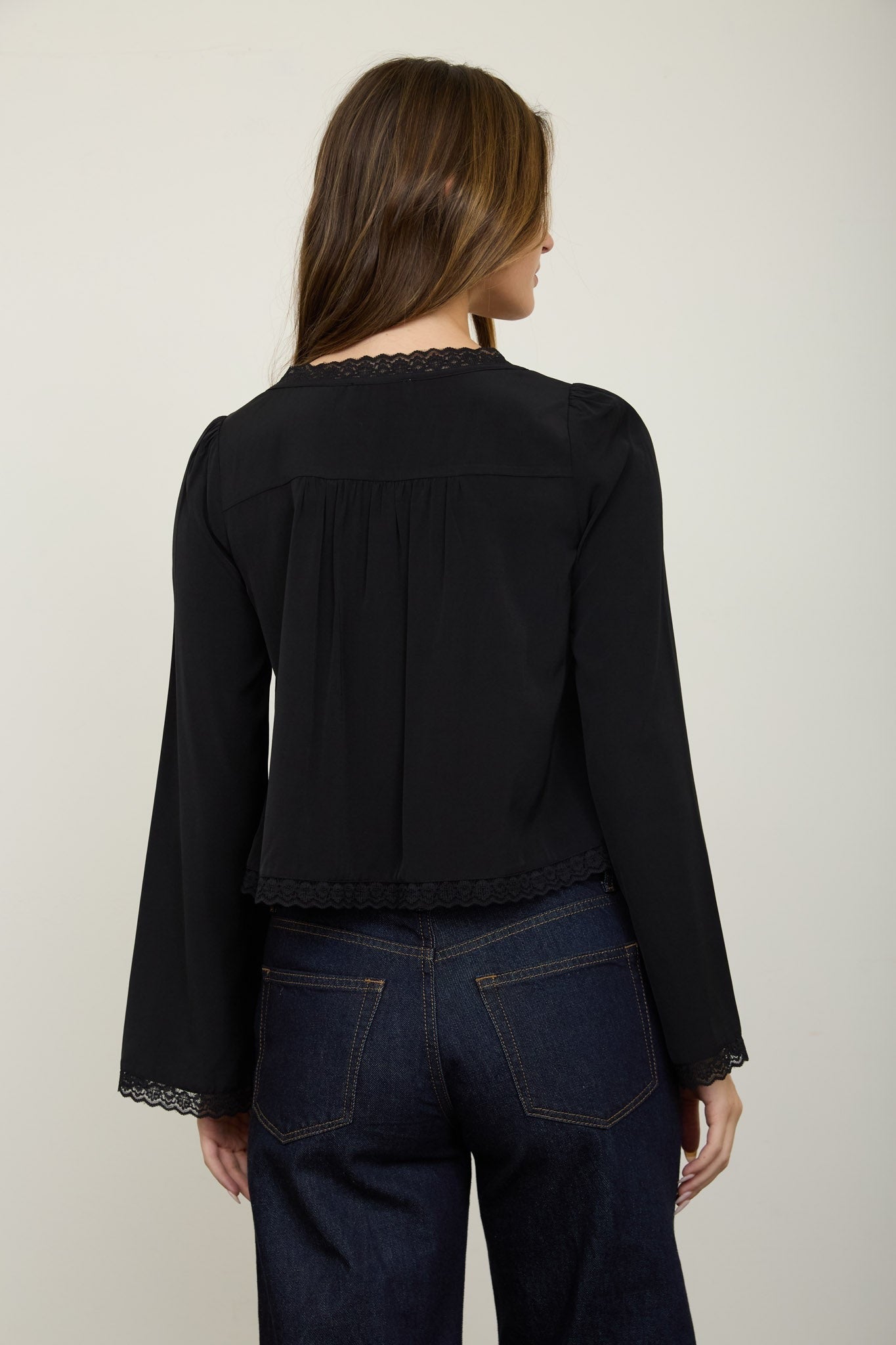 A woman shown from the back wearing a black long-sleeve blouse with delicate lace trim at the neckline and cuffs, paired with dark blue jeans, against a plain light gray studio background.