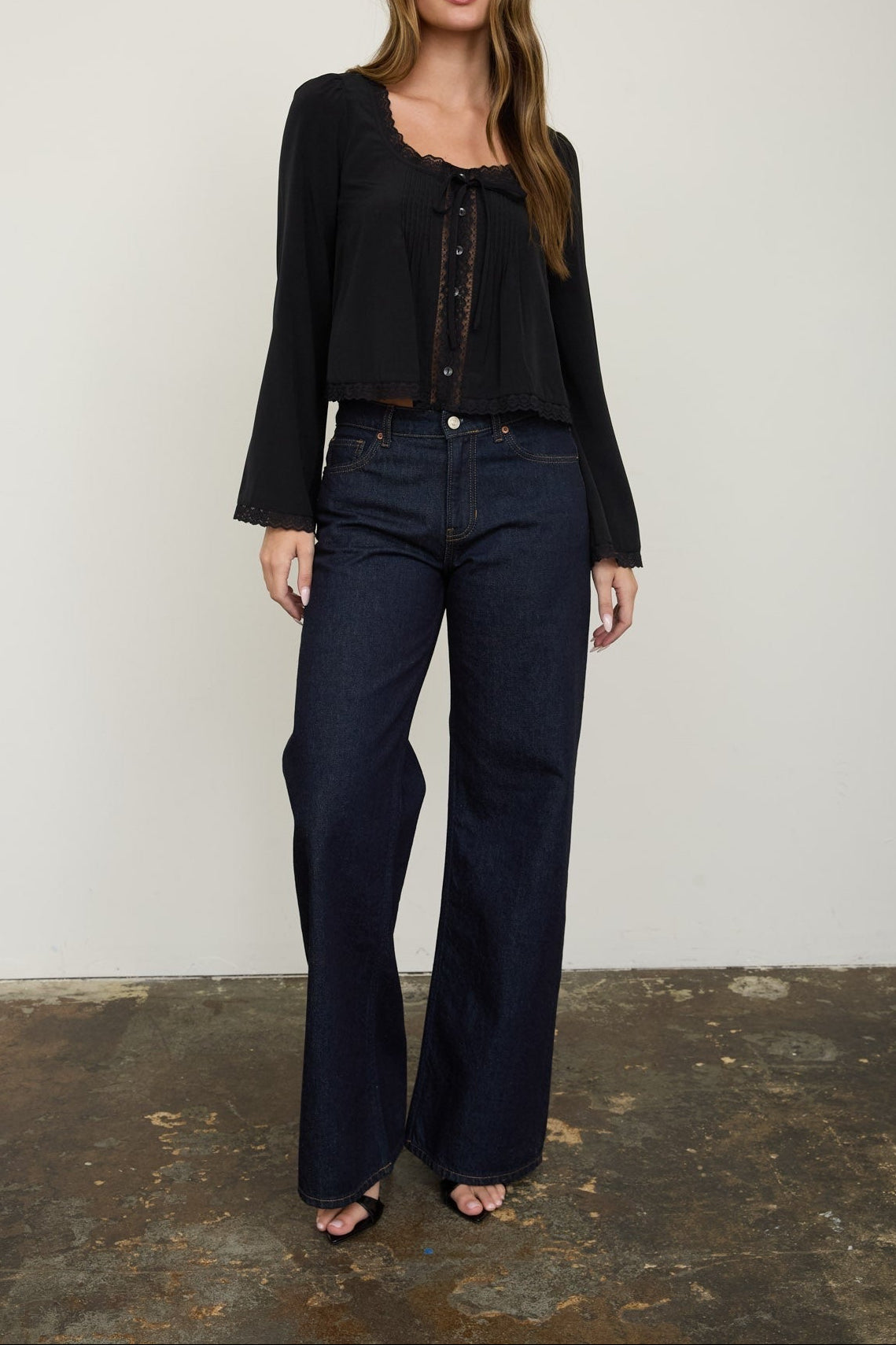 A woman modeling a black long-sleeve cropped top and dark blue wide-leg jeans against a plain off-white wall and rustic concrete floor.