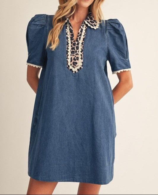 A woman wearing a blue denim-style mini dress with puff sleeves, a lace-trimmed V-neckline, and cream lace collar details, posed against a plain beige studio background.