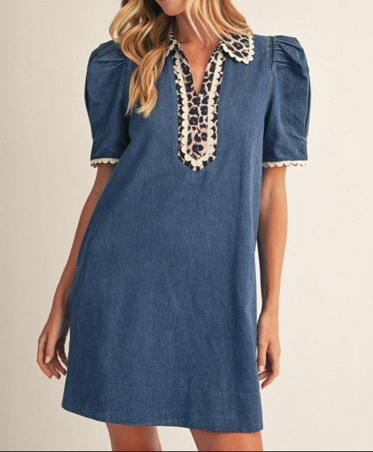 A woman wearing a blue denim-style mini dress with puff sleeves and a cream lace V-neck collar, photographed against a plain beige studio background.
