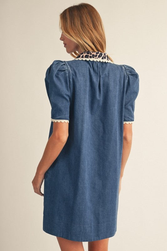 A woman wearing a blue denim dress with cream lace trim on the collar and sleeve cuffs, shown from a rear three-quarter view against a plain beige studio background.