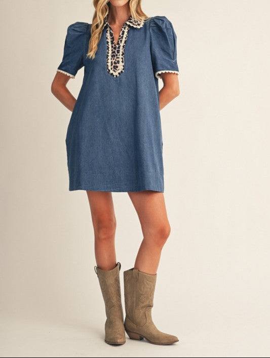 A woman modeling a short-sleeve denim dress with puff sleeves, a lace-up neckline, and white lace trim, paired with brown cowboy boots against a plain beige studio background.