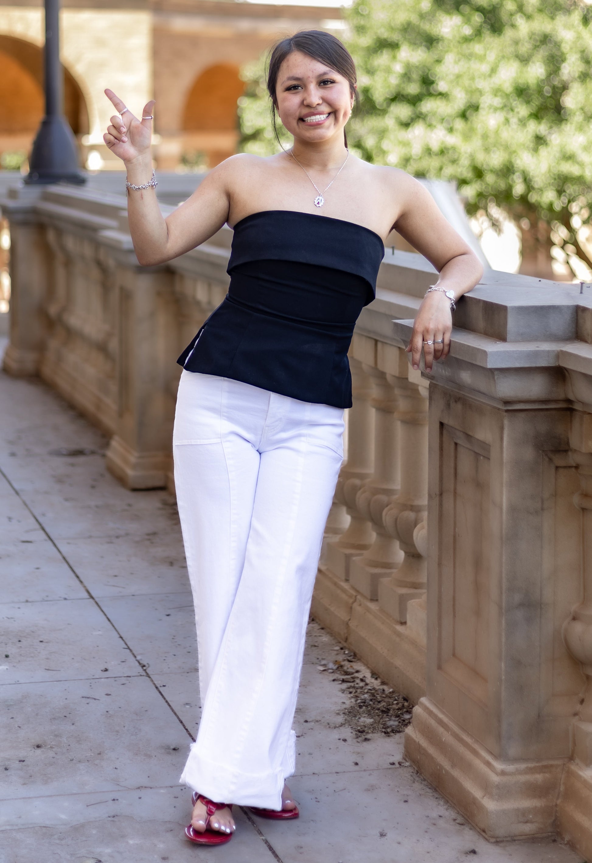 Woman in black top and white pants posing outdoors near a stone wall.