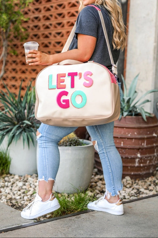 Woman holding a beige duffel bag with 'LET'S GO' text, standing outdoors.
