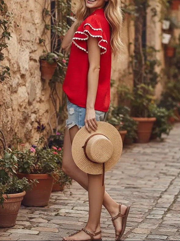 Woman in a red top and denim shorts holding a straw hat on a cobblestone street.