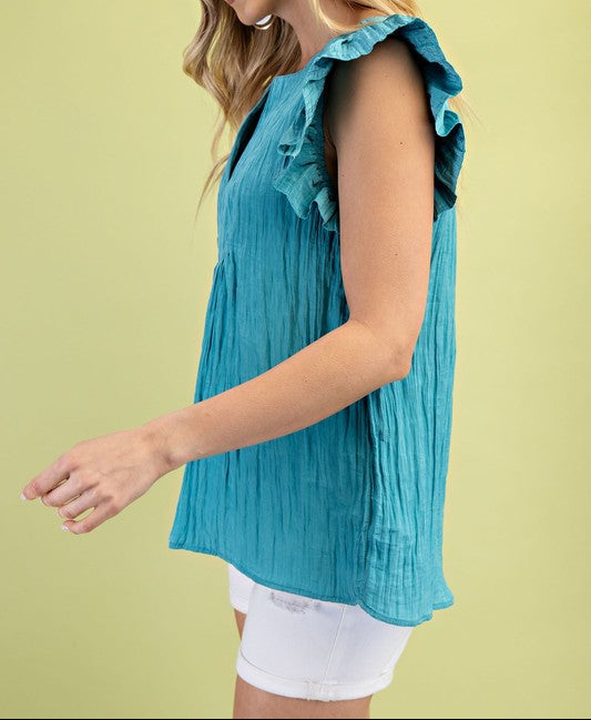 Woman wearing a teal sleeveless top with ruffled shoulders on a light green background