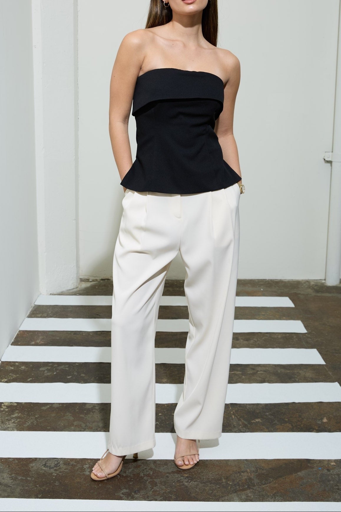 Woman wearing a black strapless top and white pants in a minimal indoor setting.