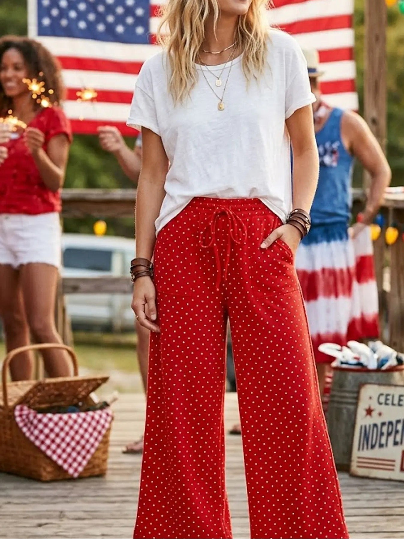 Woman in red polka dot pants and white shirt standing in front of an American flag with people around.
