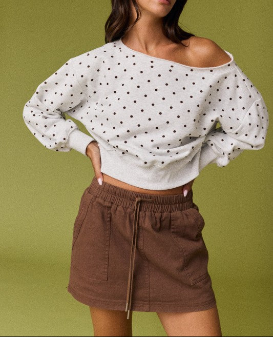A woman modeling a white off-the-shoulder long-sleeve knit top with black polka dots, paired with a brown corduroy mini skirt, against a solid olive green studio background.