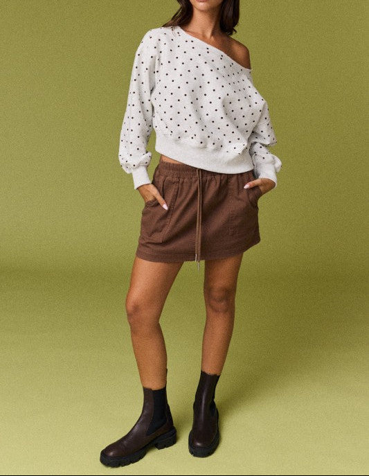 A model stands against a solid olive green studio background wearing a white long-sleeve top with black polka dots, a brown mini skirt, and black ankle boots.