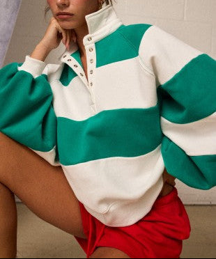 A woman wearing a green and white striped quarter-zip sweatshirt with balloon sleeves, paired with red shorts, posing in front of a light-colored brick wall.