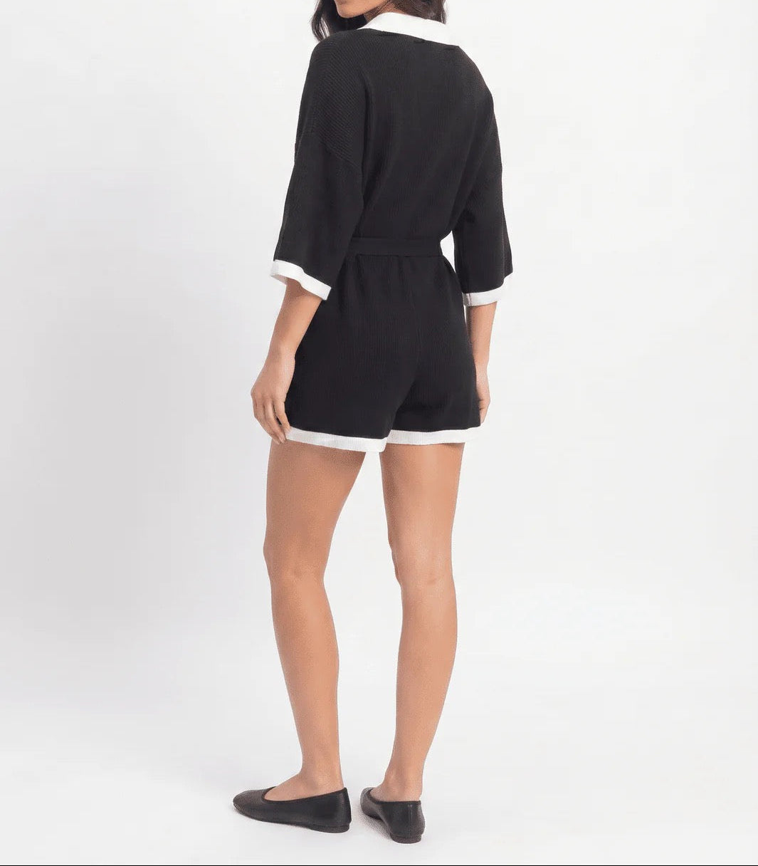A woman modeling a black short romper with white contrast trim on the collar, cuffs, and hem, shown from a rear three-quarter angle against a plain white studio background.
