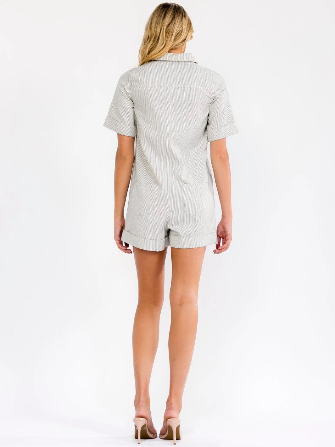 A woman viewed from the back wearing a light gray short-sleeve collared romper with matching shorts, paired with beige high heels, against a plain white studio background.