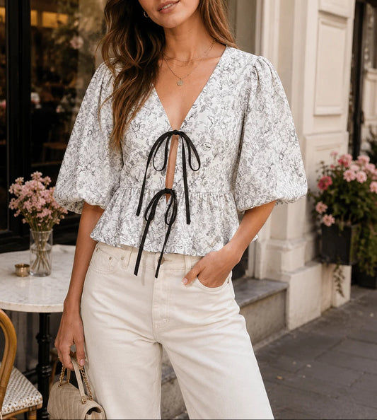 A woman wearing a white short-sleeve blouse with a black floral pattern, a black tie detail at the V-neck, and white pants, standing on a city street.