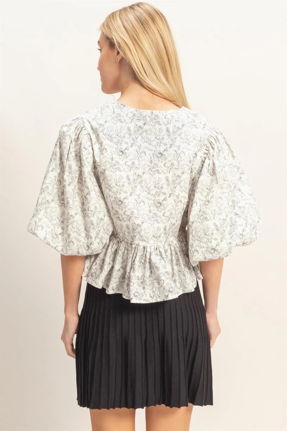 A woman shown from the back wearing a white floral puff-sleeve top with a gathered peplum hem and a black pleated mini skirt, against a plain light beige studio background.