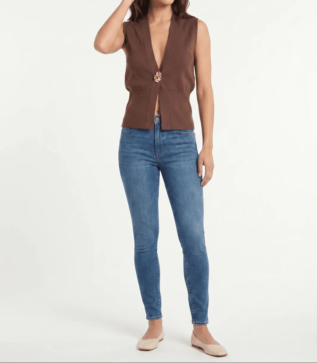 A woman modeling a sleeveless brown vest with a deep V-neck and a decorative gold-toned clasp, paired with blue skinny jeans and beige flats, against a plain white studio background.