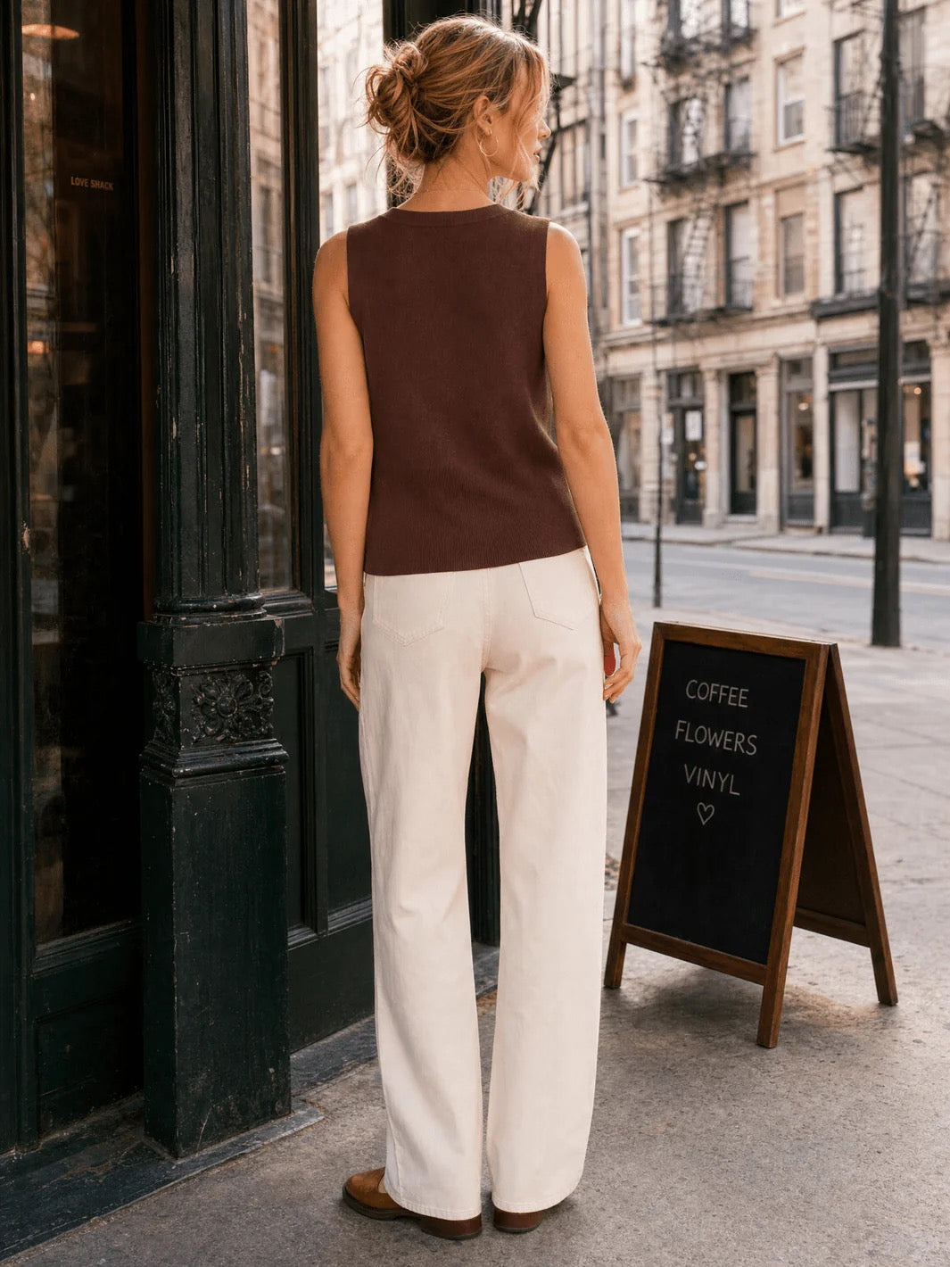 A woman stands on a city sidewalk facing a storefront, wearing a sleeveless dark brown knit top and off-white wide-leg pants. A small chalkboard sign reading 'COFFEE FLOWERS VINYL' sits beside her.