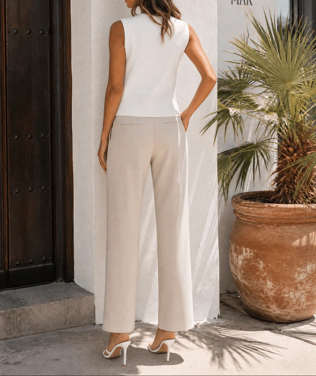 Woman in a white sleeveless top and beige pants standing against a white wall with a plant in the background.