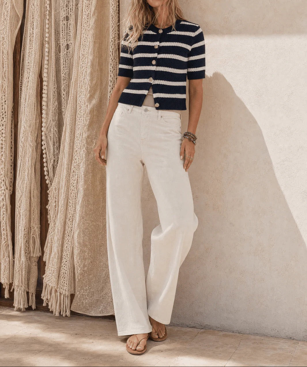 A woman stands against a textured beige wall wearing a navy and white striped short-sleeve button-front top with white wide-leg pants and tan sandals. A cream woven textile hangs on the left side of the frame.