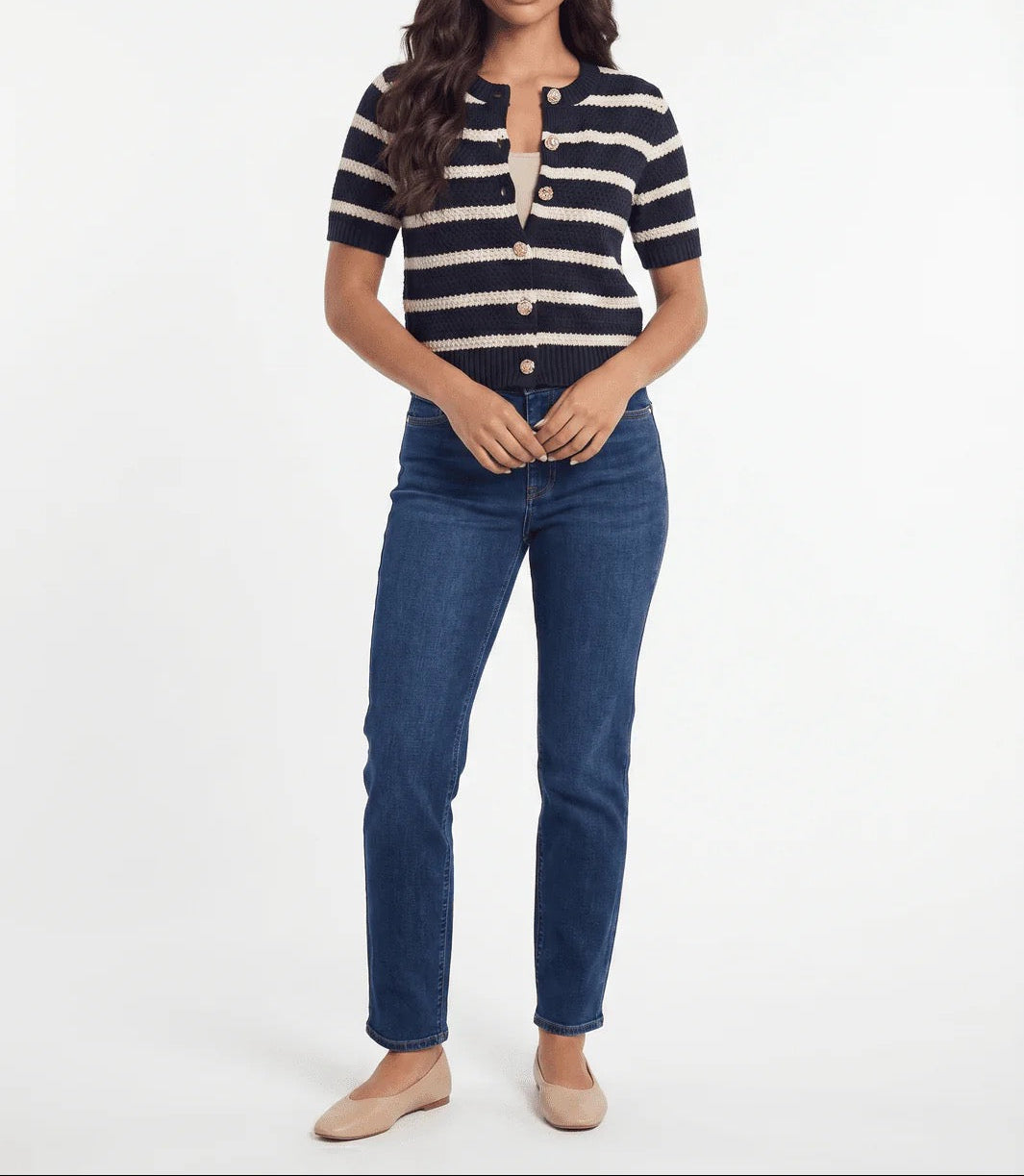 A woman modeling a short-sleeve knit top with navy and cream horizontal stripes, paired with dark blue jeans and beige flats, against a plain white studio background.