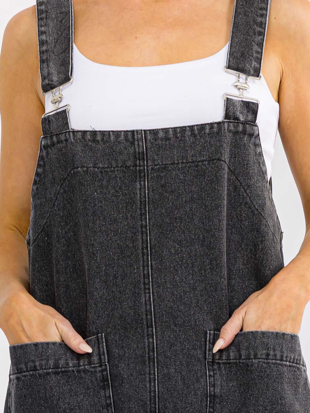 Close-up of a person wearing dark gray washed denim overall with adjustable shoulder straps and front patch pockets, layered over a white tank top.