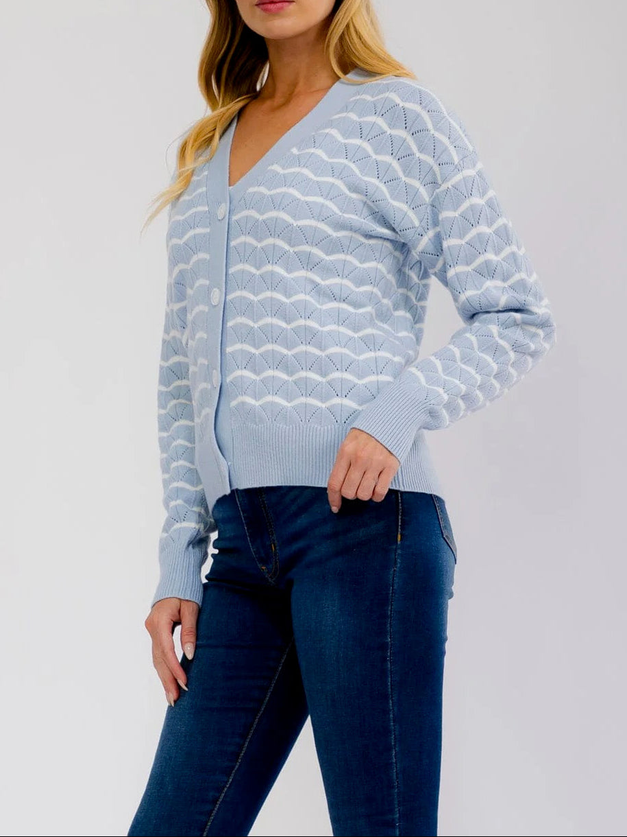 A woman modeling a light blue and white wavy patterned knit cardigan sweater with dark blue jeans, photographed against a plain light gray studio background.