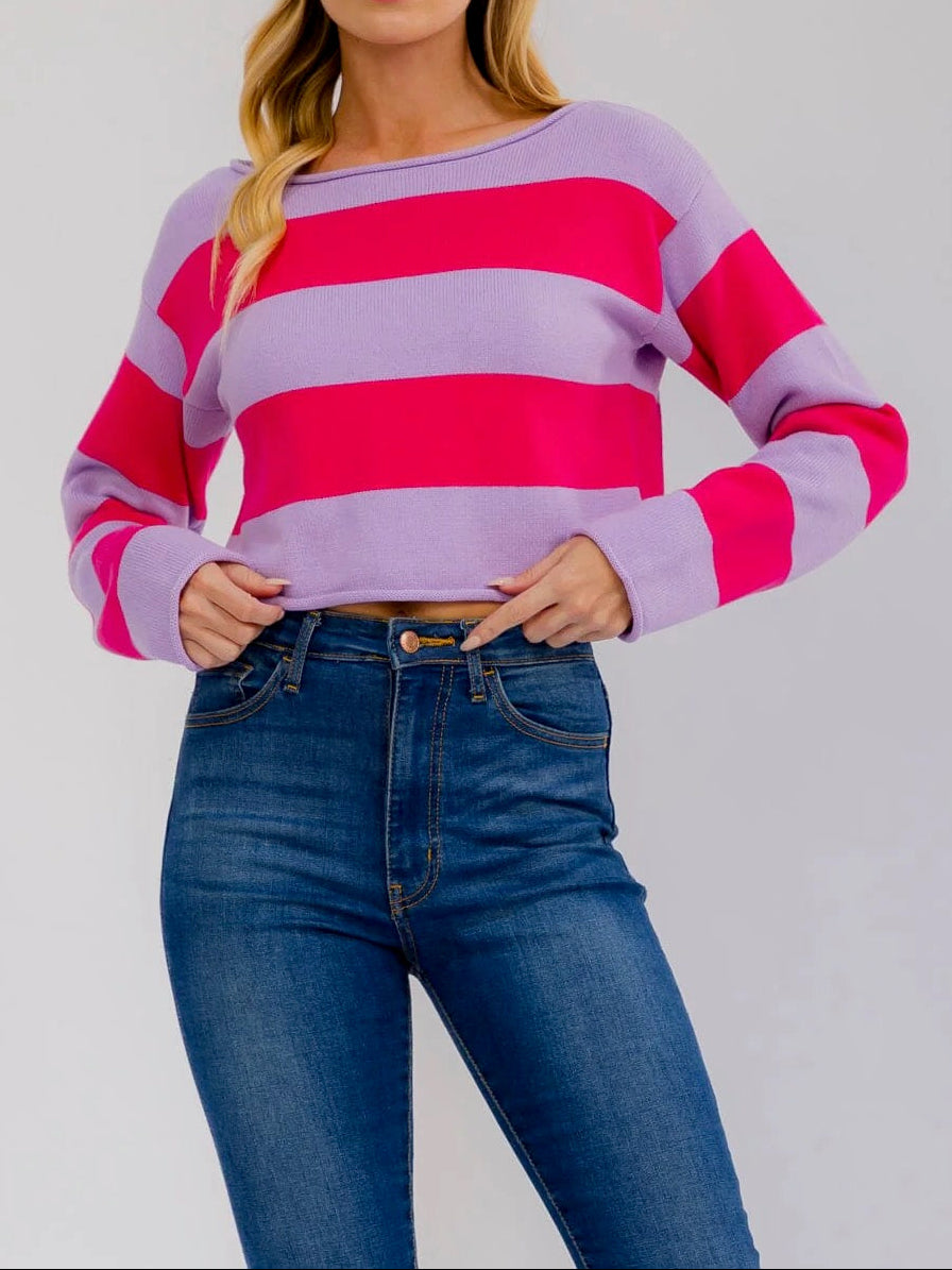 A woman modeling a long-sleeve knit sweater with bold horizontal stripes in lavender purple and bright pink, paired with dark blue jeans against a light gray studio background.