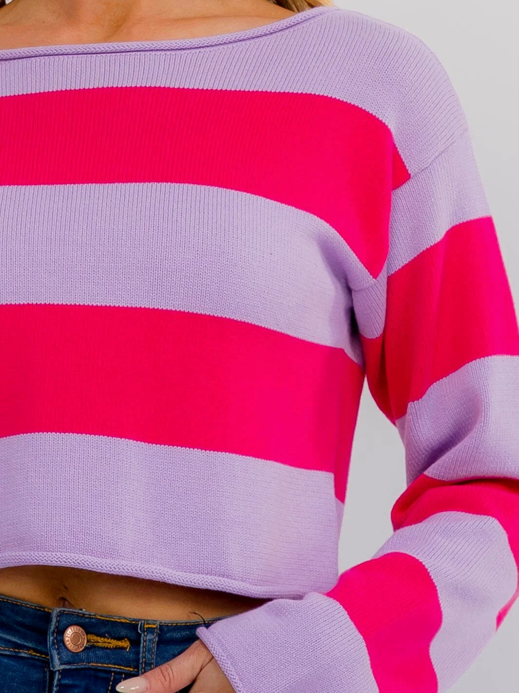A close-up of a person wearing a cropped knit sweater with wide horizontal stripes in light lavender and bright pink, paired with blue jeans.