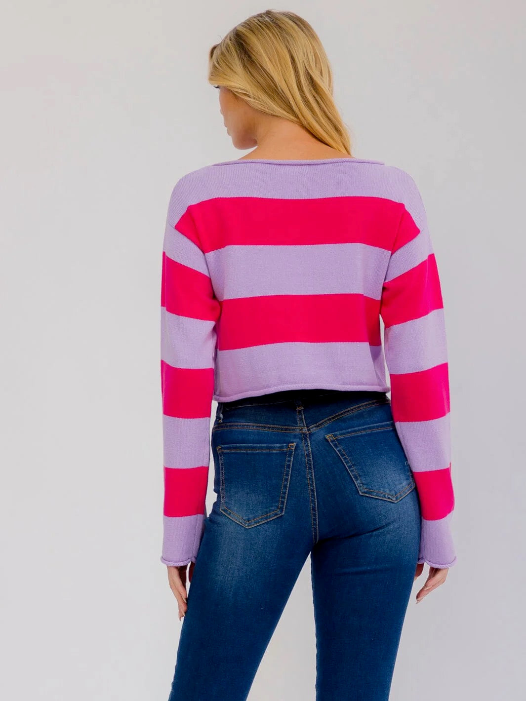 A woman viewed from the back wearing a cropped long-sleeve knit top with bold horizontal pink and lavender stripes, paired with dark blue jeans, against a light gray studio background.