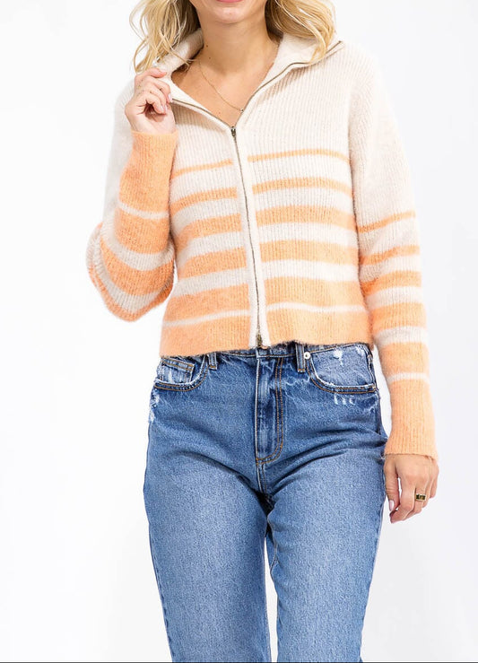 A woman wearing a fuzzy knit zip-up hoodie with horizontal peach and cream stripes, paired with blue jeans, photographed against a plain white background.