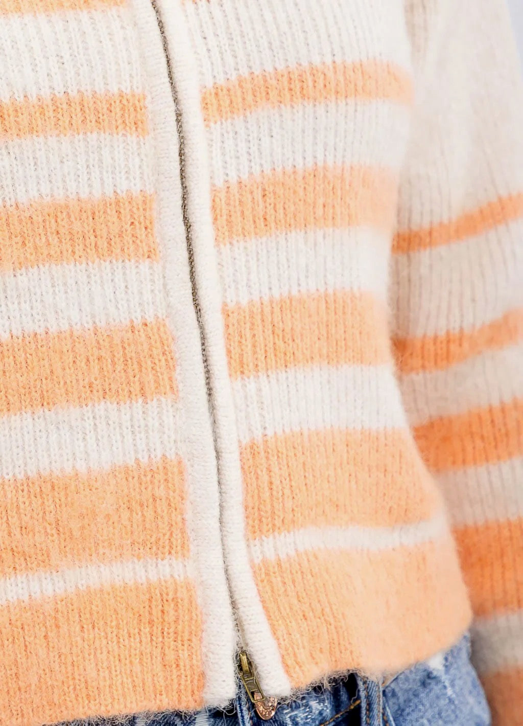 Close-up of a fuzzy knit zip-up cardigan with horizontal peach and off-white stripes, shown worn with blue jeans.