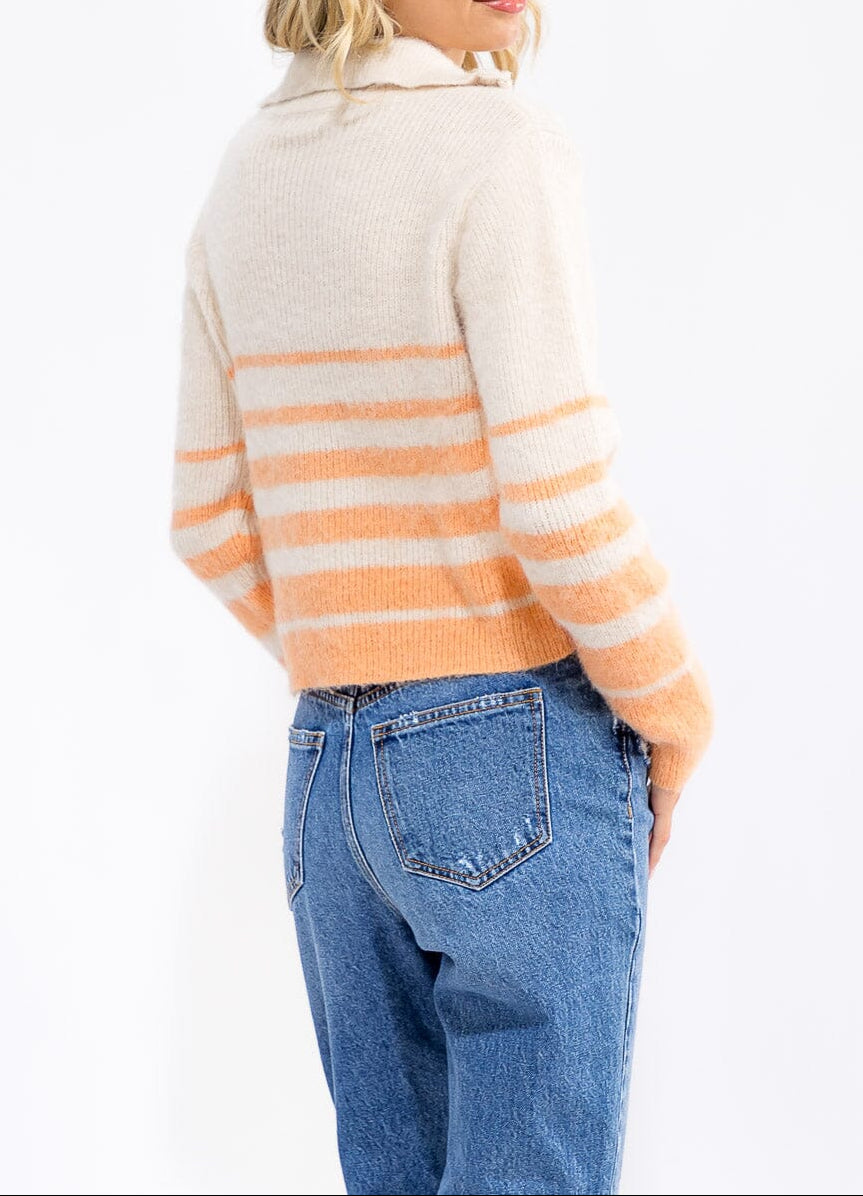 A woman shown from the back wearing a cream knit sweater with horizontal peach-orange stripes and blue jeans, against a plain white studio background.