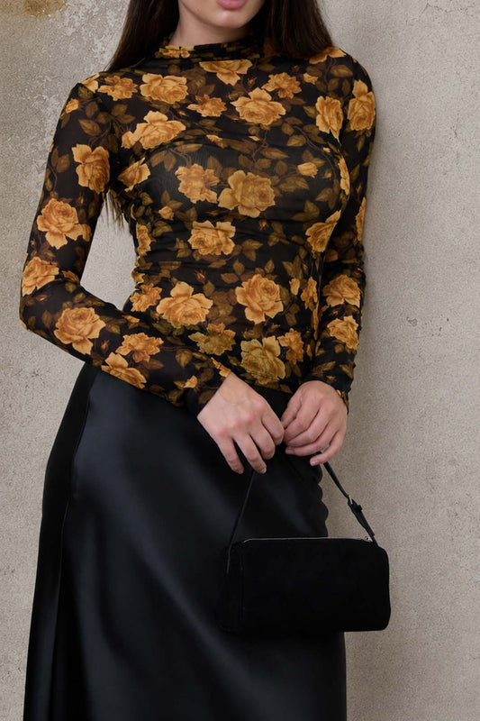Woman wearing a black floral top and black skirt holding a black handbag against a textured beige wall.