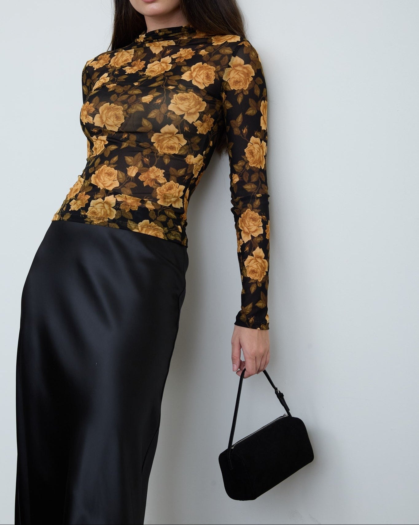 A woman wearing a sheer black long-sleeve top with a mustard yellow rose pattern, paired with a black satin skirt, holding a small black handbag against a plain light gray wall.