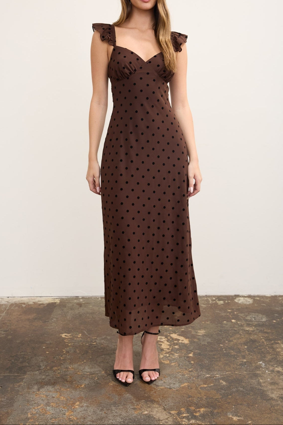 A woman modeling a dark brown midi dress with a black polka dot pattern, ruffled shoulder straps, and a sweetheart neckline, standing against a plain off-white wall.