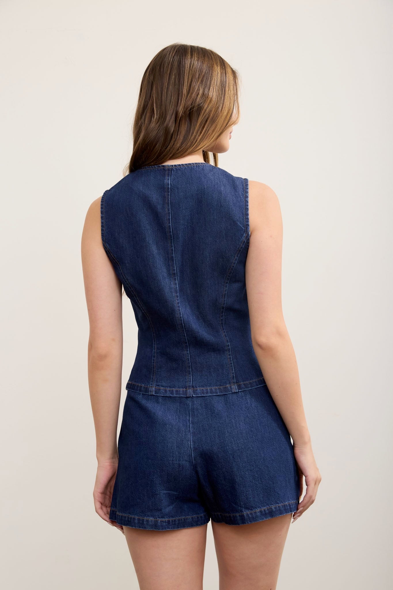 A woman viewed from the back wearing a sleeveless dark blue denim romper against a plain light beige studio background.