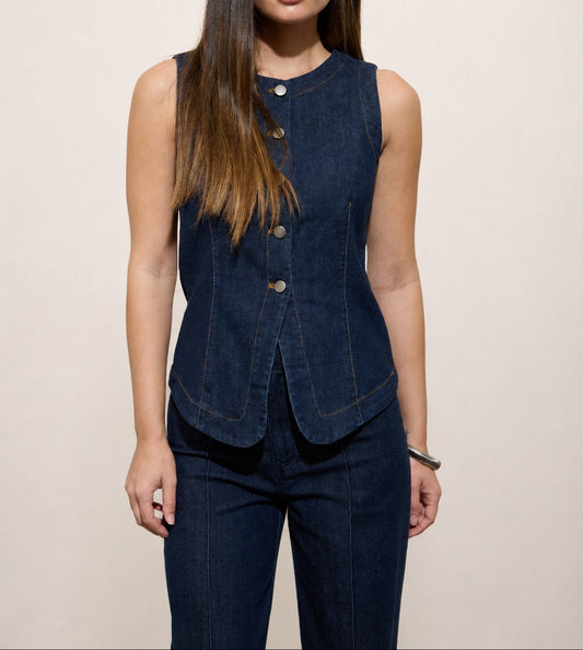 A woman modeling a dark blue denim sleeveless vest with front buttons, paired with matching dark blue denim pants, against a plain beige studio background.