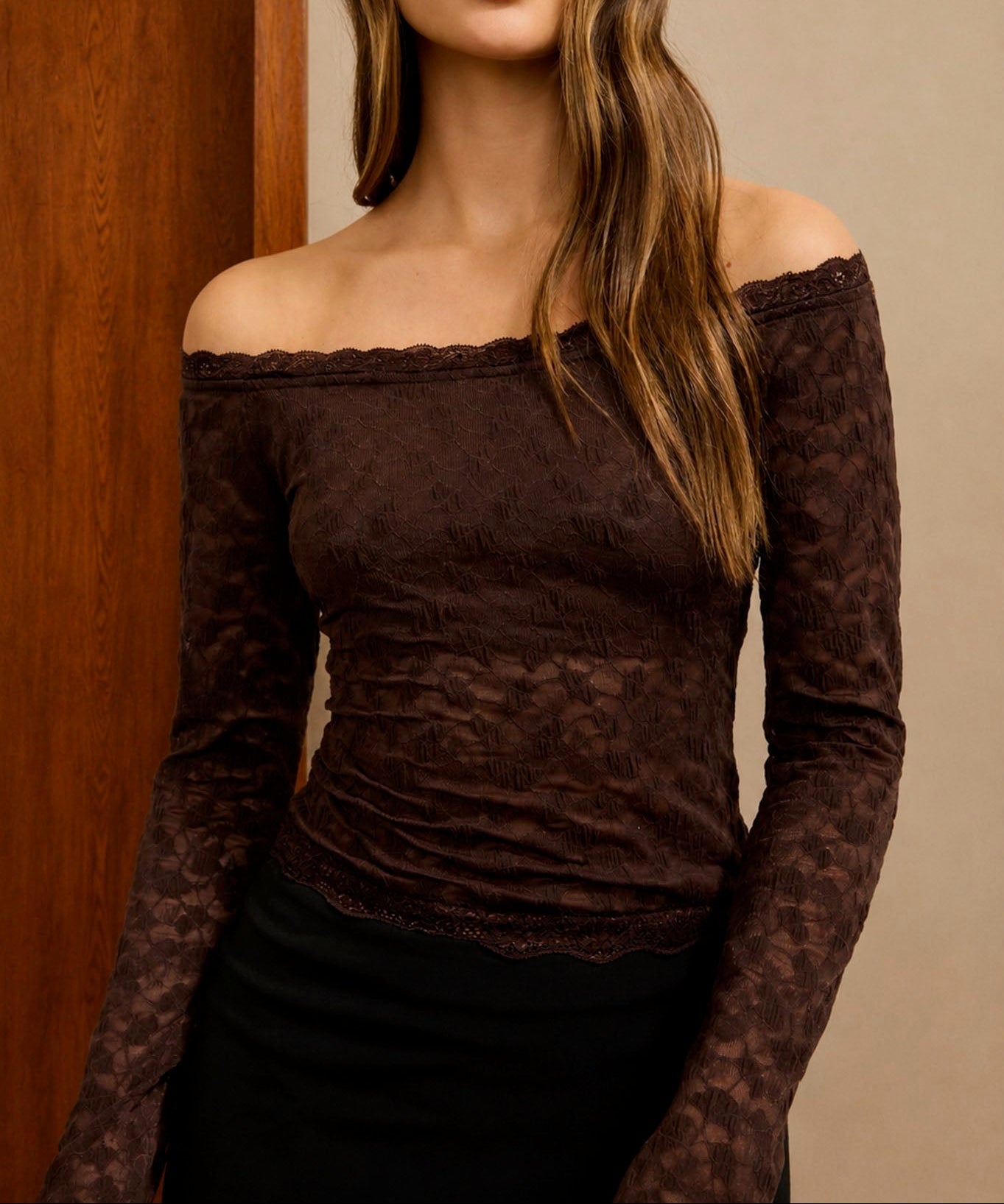 A woman wearing a dark brown off-the-shoulder lace long-sleeve top and a black skirt, standing in front of a beige wall and wooden door frame.