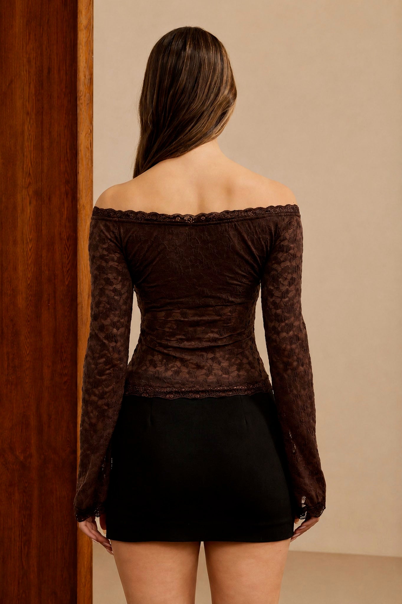 Woman wearing a brown off-shoulder top with lace details and a black skirt against a beige wall.