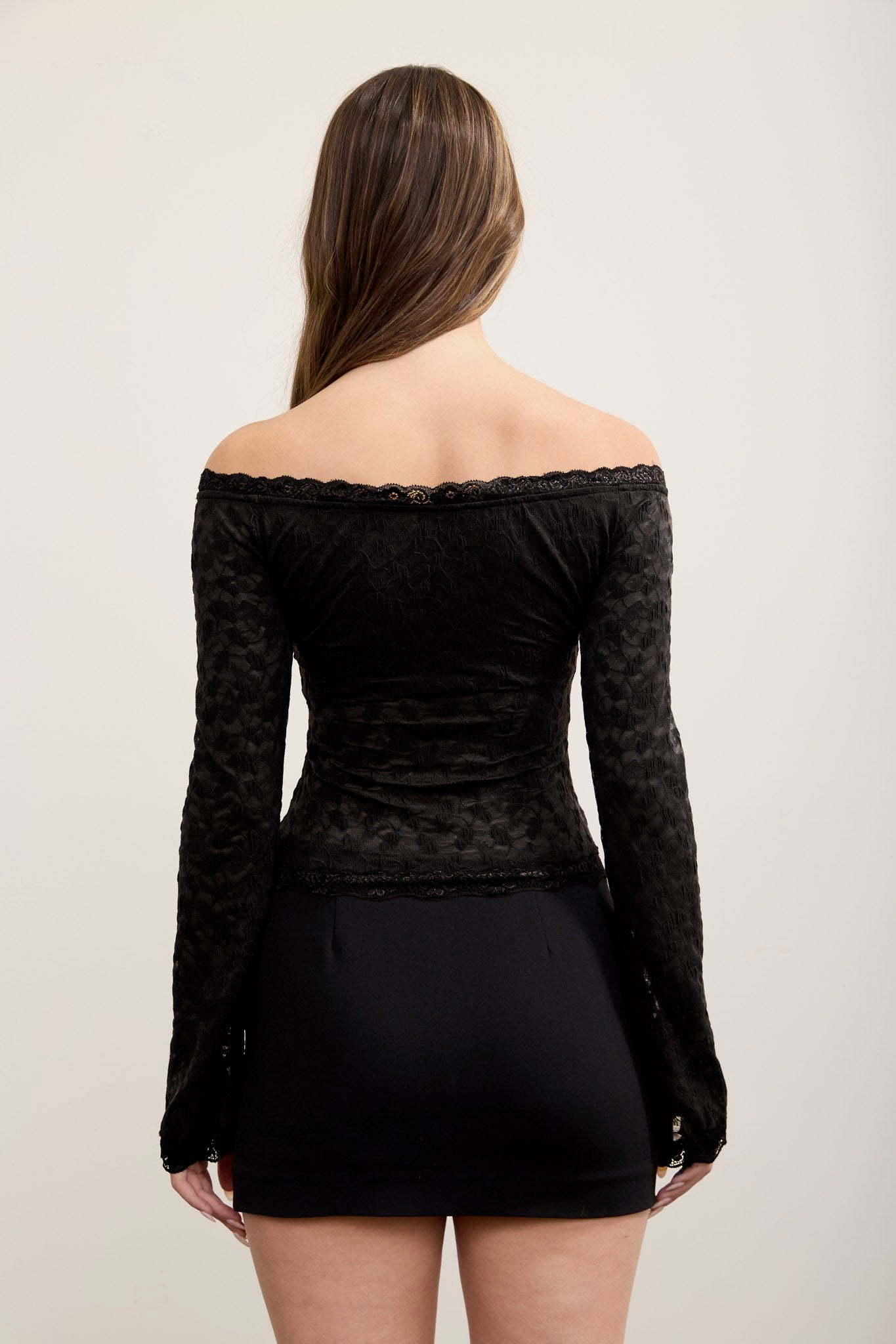 A woman viewed from the back wearing a black off-the-shoulder long-sleeve lace top and a black mini skirt against a plain light background.