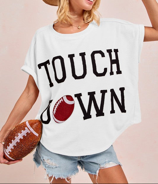 A woman wearing a white oversized graphic t-shirt with 'TOUCH DOWN' printed in black and a football graphic, styled with a brown cowboy hat, denim shorts, and holding a football against a solid beige background.