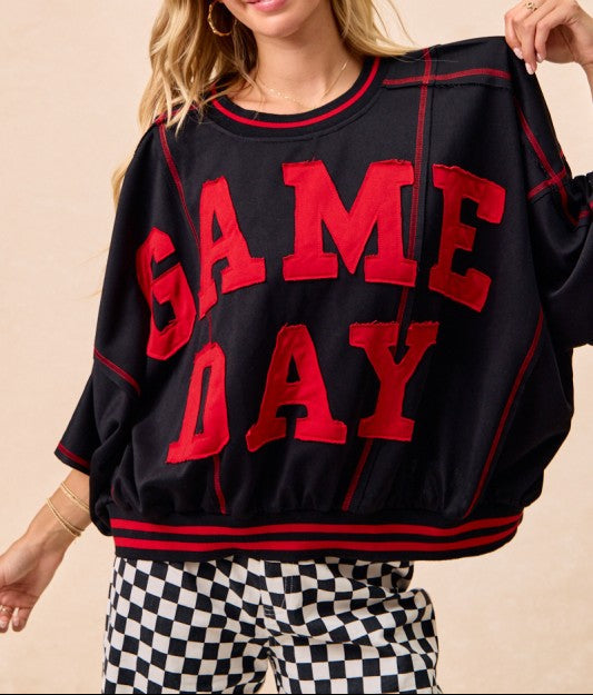 A woman wearing a black crewneck sweatshirt with 'SAME DAY' printed in red varsity-style letters across the front, paired with black-and-white checkered pants, against a plain beige background.