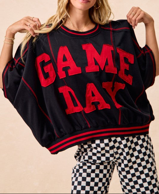 A woman wearing a black bubble-sleeve sweatshirt with 'GAME DAY' in red varsity letters, paired with black-and-white checkered pants, against a warm beige background.