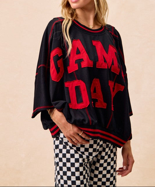 A woman wearing a black oversized graphic sweatshirt with bold red 'GAME DAY' lettering on the front, paired with black-and-white checkered pants, against a plain beige studio background.