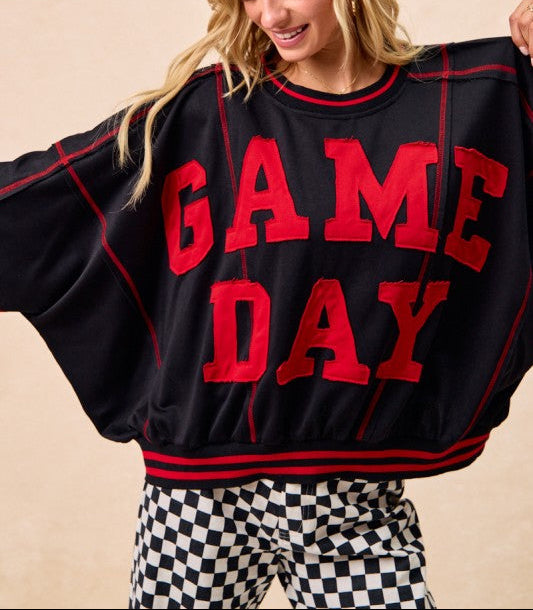A woman wearing a black oversized sweatshirt with 'GAME DAY' in large red letters, paired with black-and-white checkered pants, against a plain beige background.