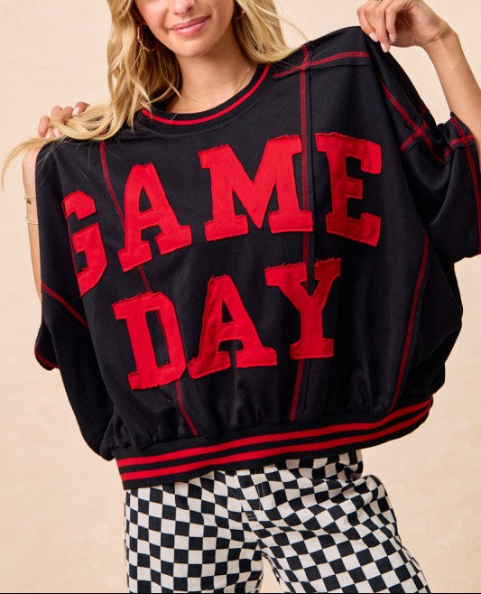 A woman modeling a black crewneck sweatshirt with 'GAME DAY' printed in bold red letters across the front, paired with black and white checkered pants against a warm beige background.