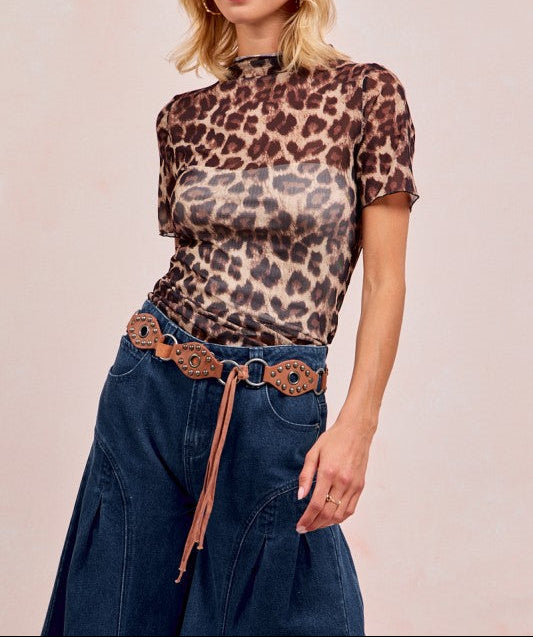 A woman modeling a short-sleeve leopard-print mock neck top with a sheer mesh texture, paired with a brown woven belt and dark blue denim skirt, against a solid light beige background.